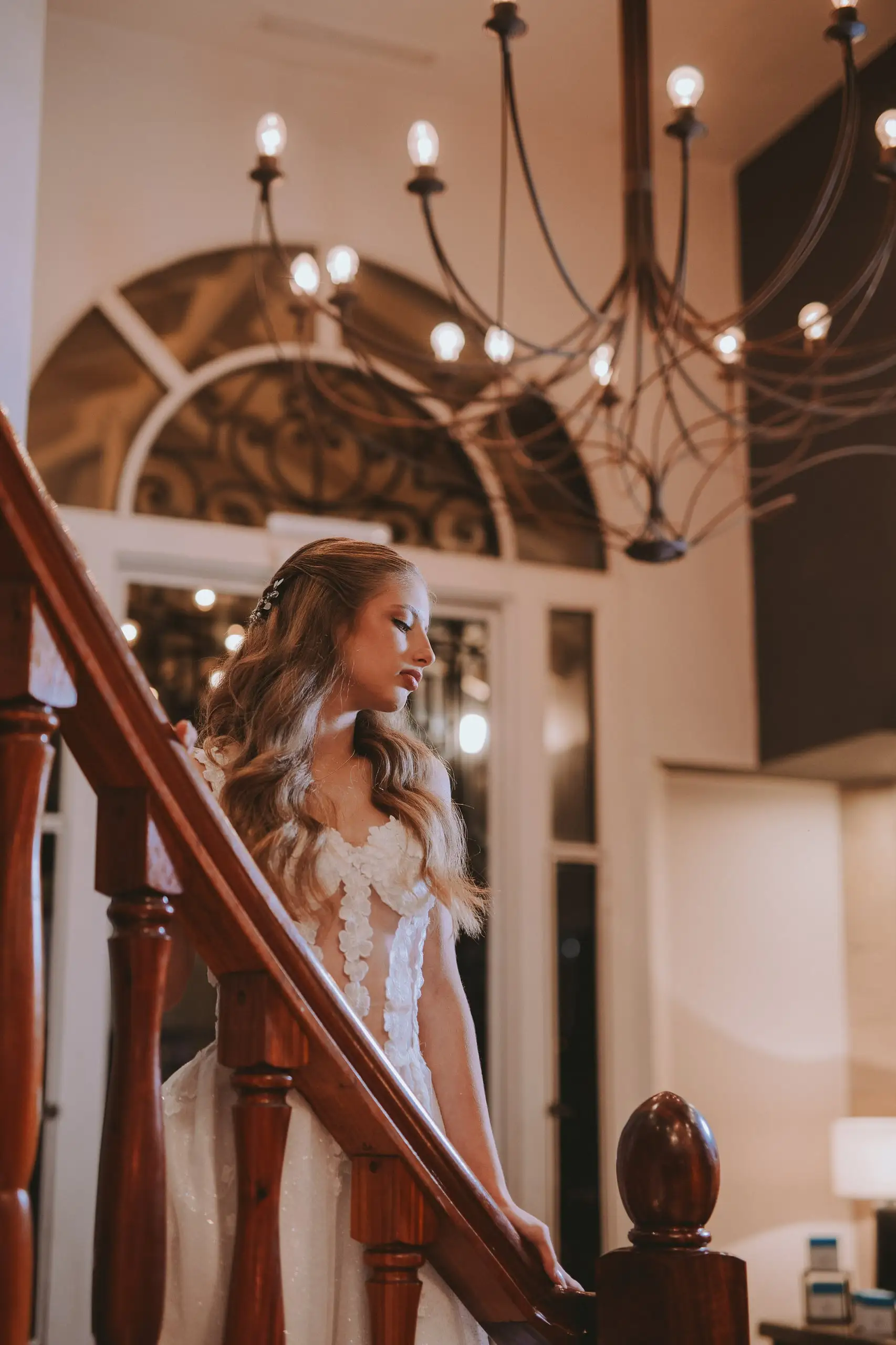 La quinceañera baja por una escalera de madera bajo una araña de luces cálidas.