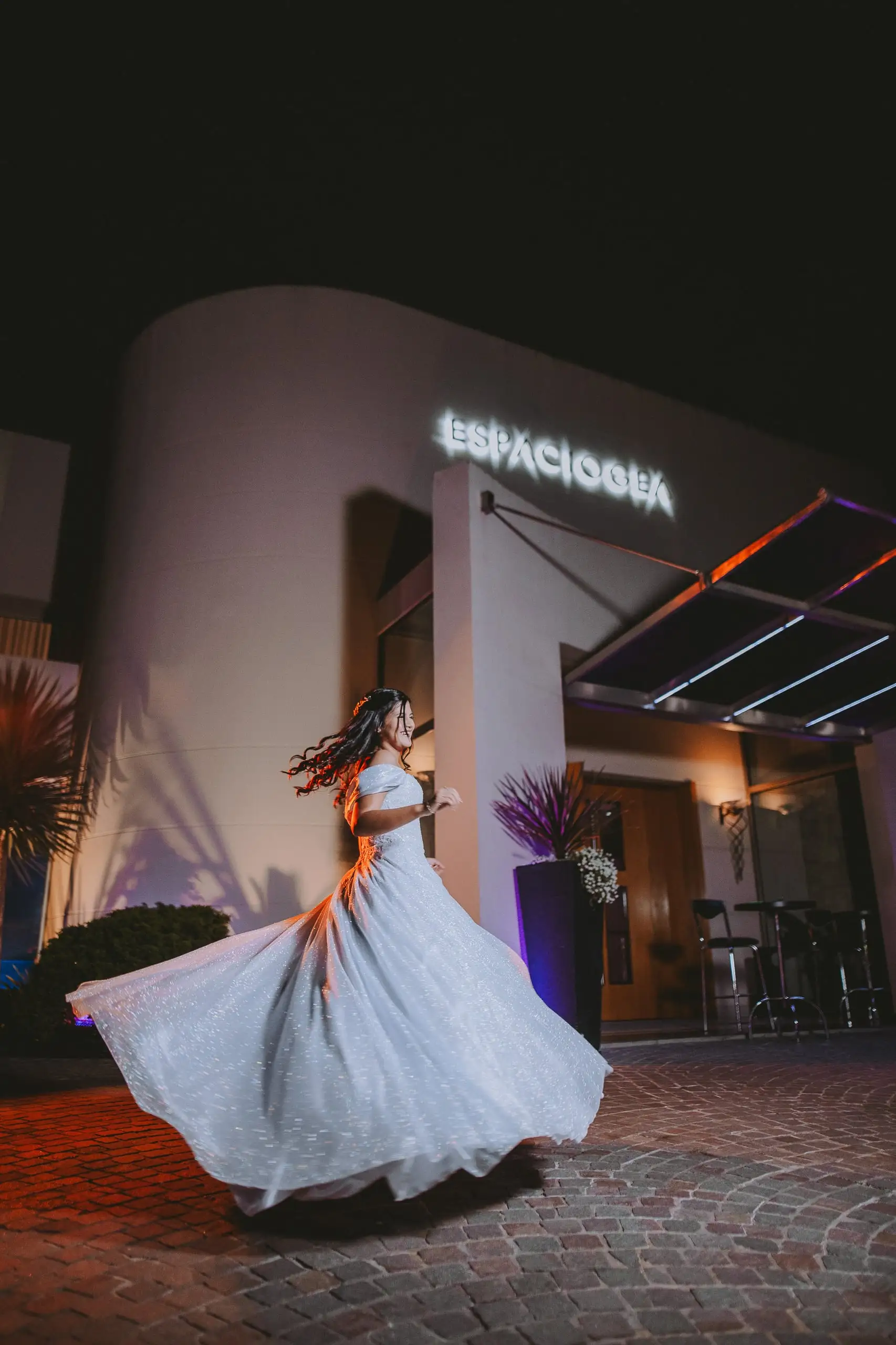 La quinceañera gira y hace volar su vestido en la entrada del salón, de noche.