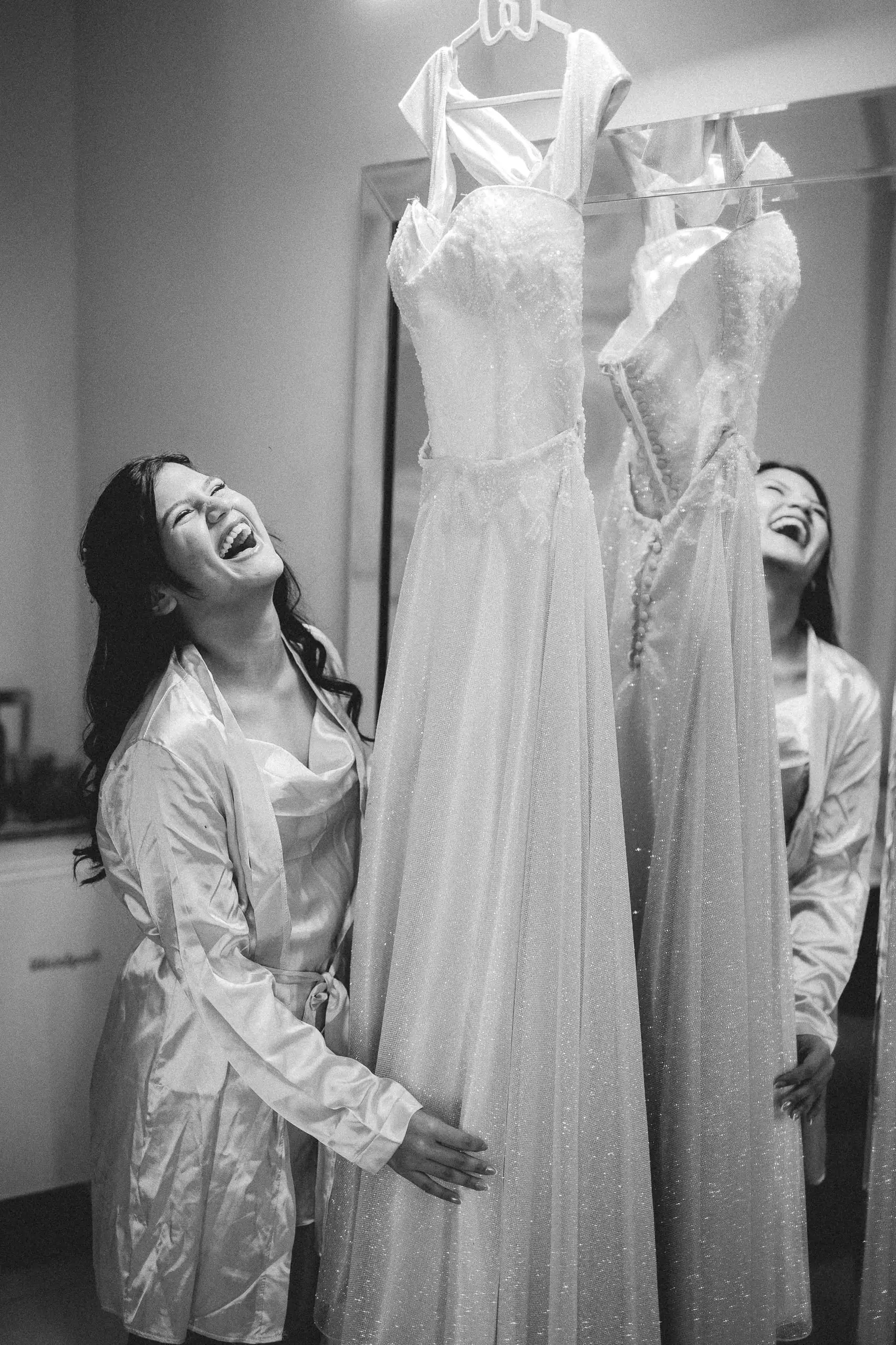 Durante la preparación, la quinceañera ríe en bata junto a sus vestidos colgados. En blanco y negro.