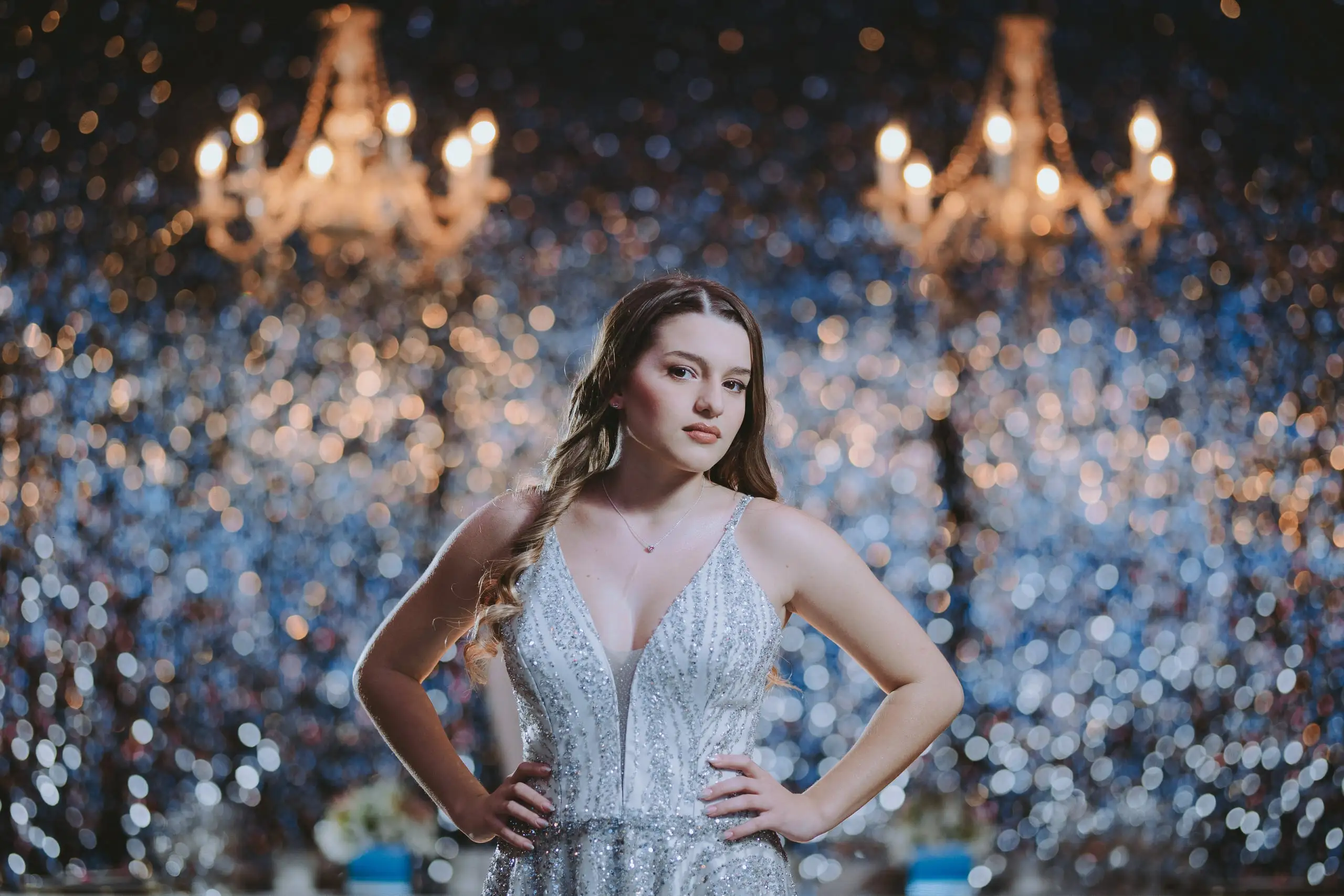 La quinceañera posa con las manos en la cintura frente a una pared de luces y arañas de cristal desenfocadas.