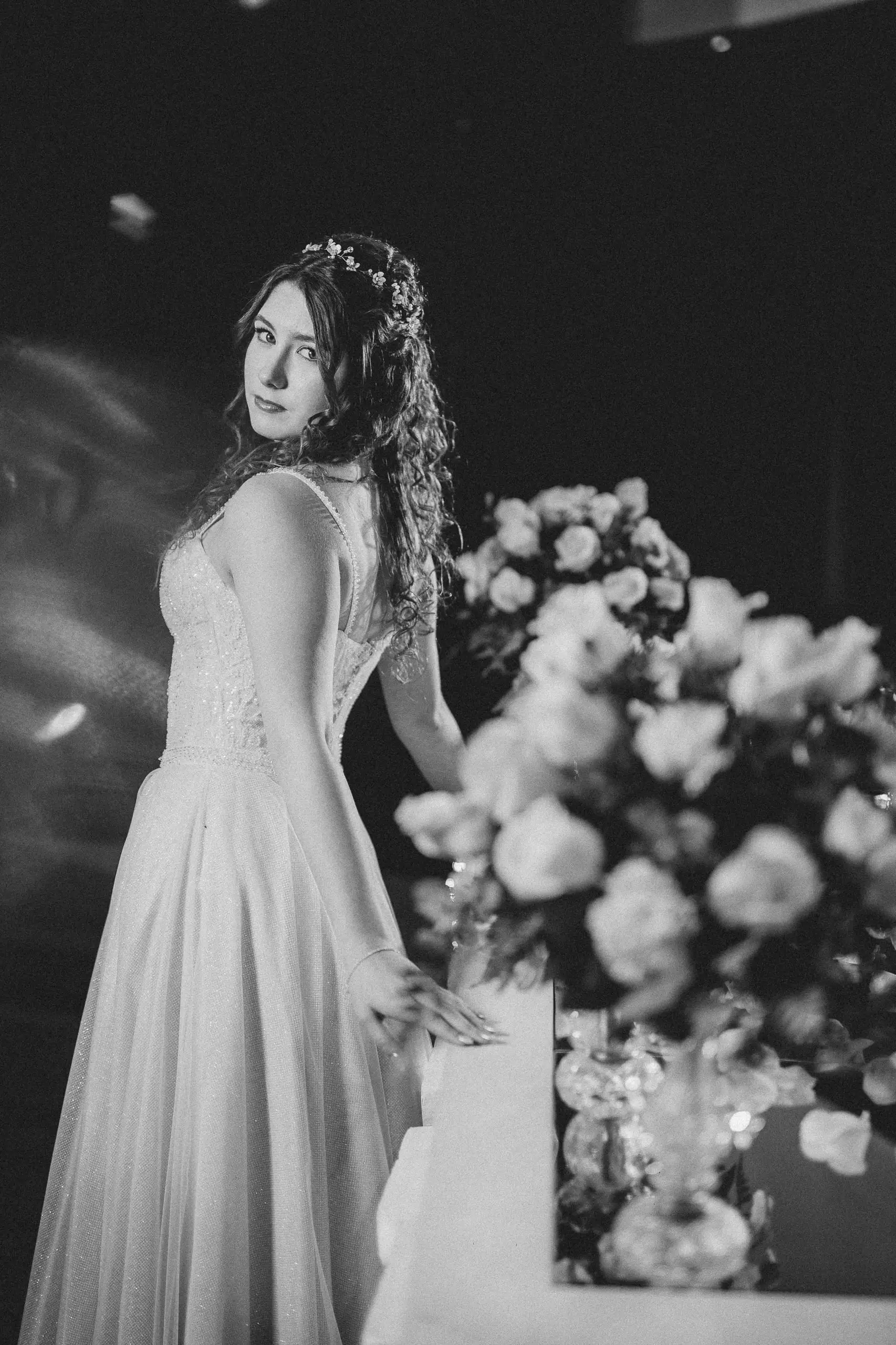 En blanco y negro, la quinceañera mira por sobre el hombro junto a un arreglo de rosas blancas.