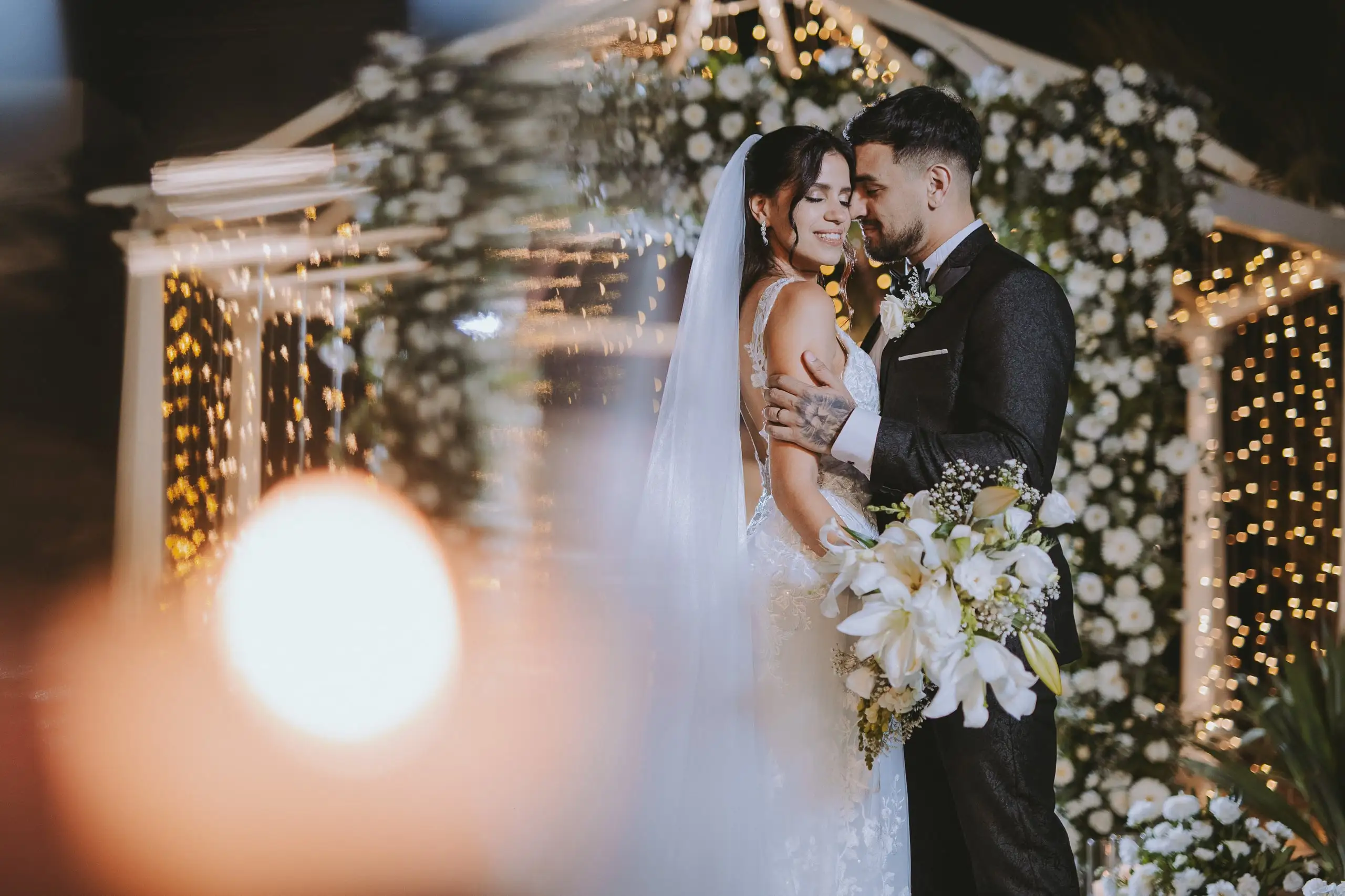 Los novios abrazados bajo el gazebo florido de noche, con luces cálidas desenfocadas en primer plano.
