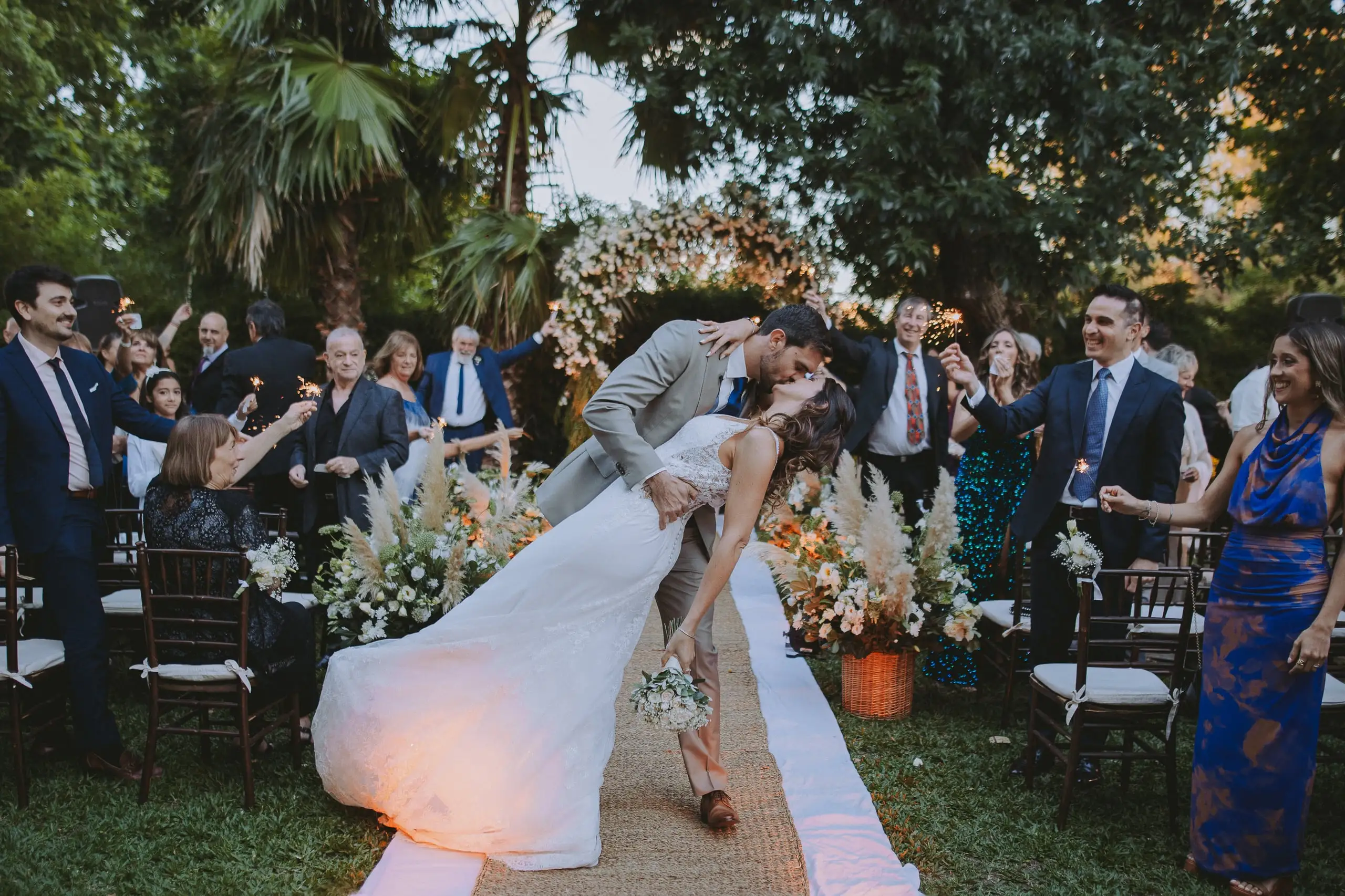 Al terminar la ceremonia al aire libre, el novio inclina a la novia para besarla en el pasillo mientras los invitados festejan.