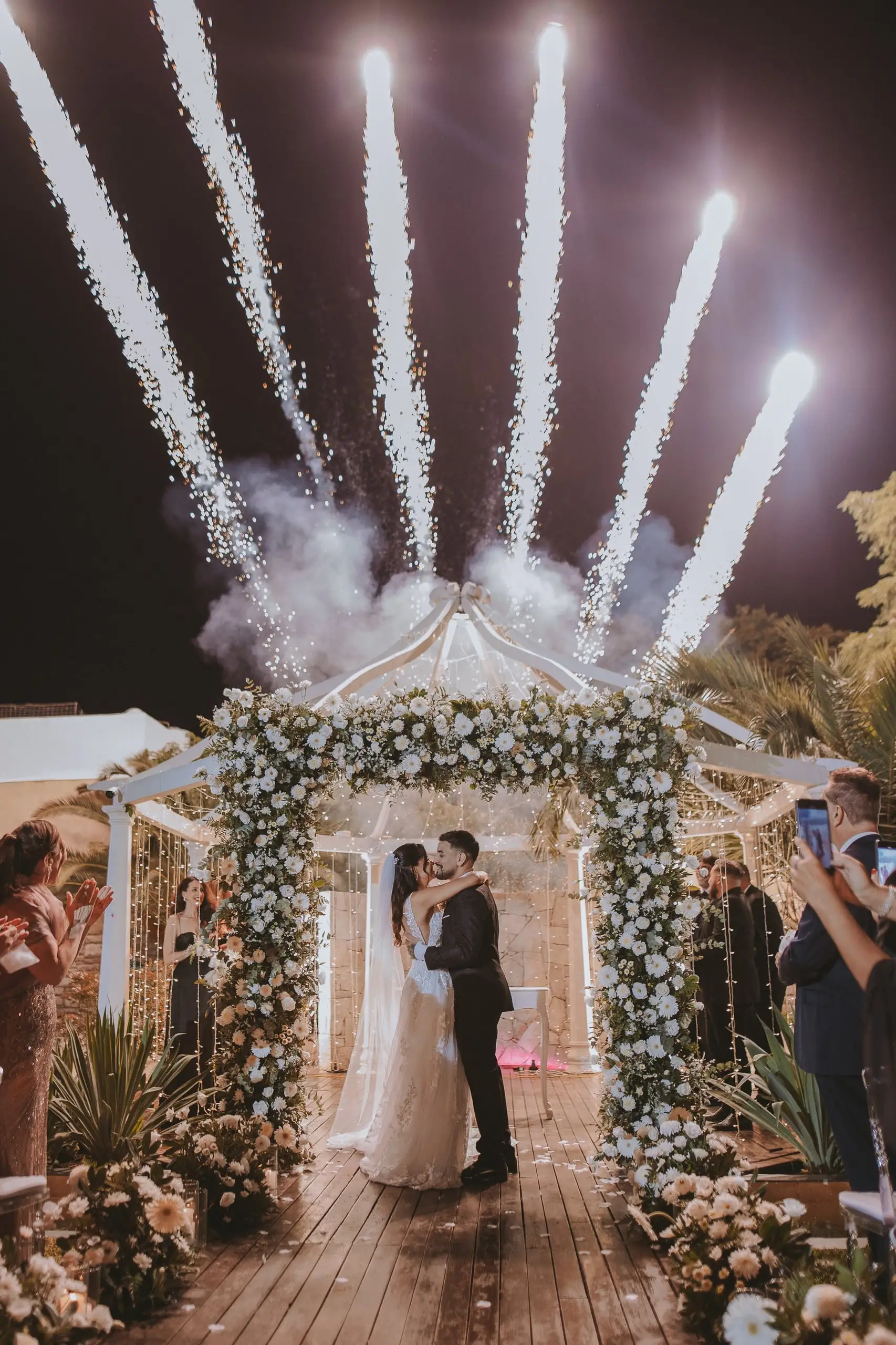 Los novios se besan bajo un gazebo cubierto de flores blancas mientras se encienden fuegos de luz fría sobre ellos.