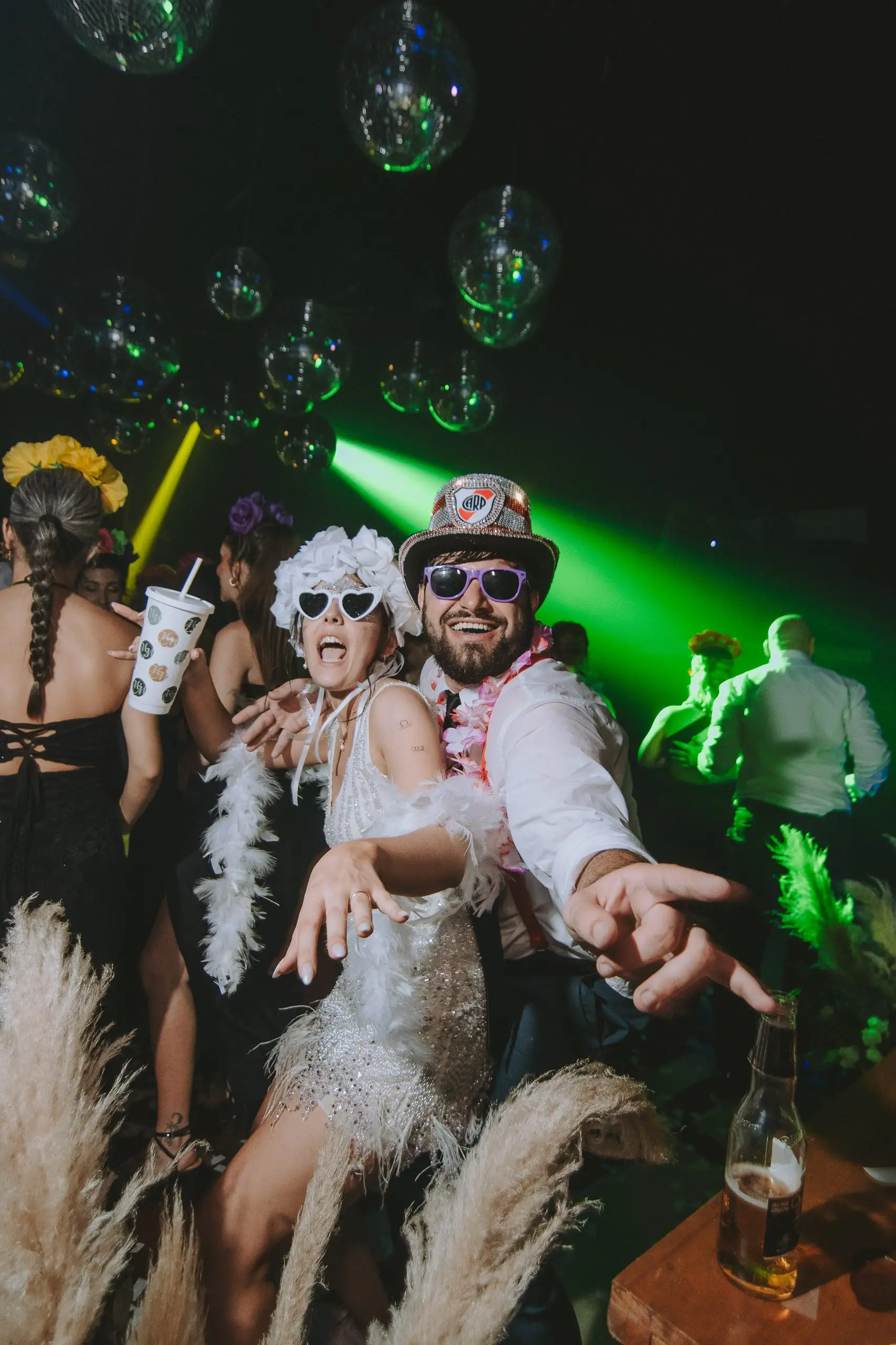 Dos invitados con anteojos de cotillón, galera y boa de plumas bailan en la fiesta entre luces verdes.