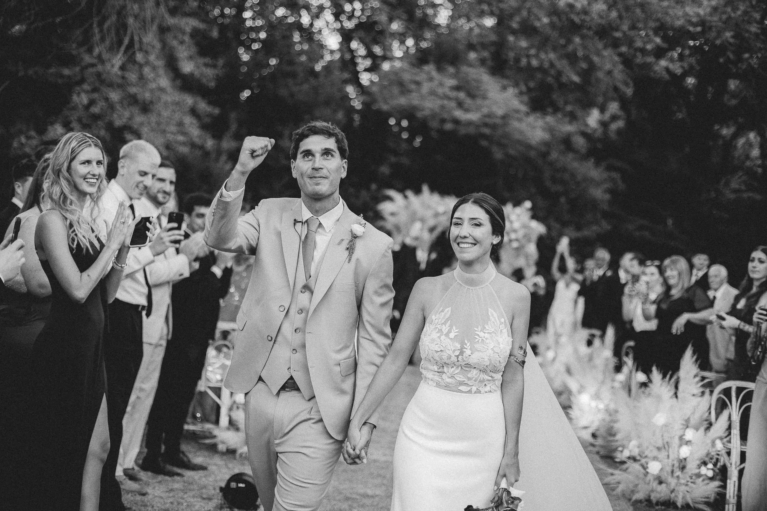 Maqui y Nico salen de la ceremonia de la mano; él levanta el puño entre los aplausos de los invitados. En blanco y negro.