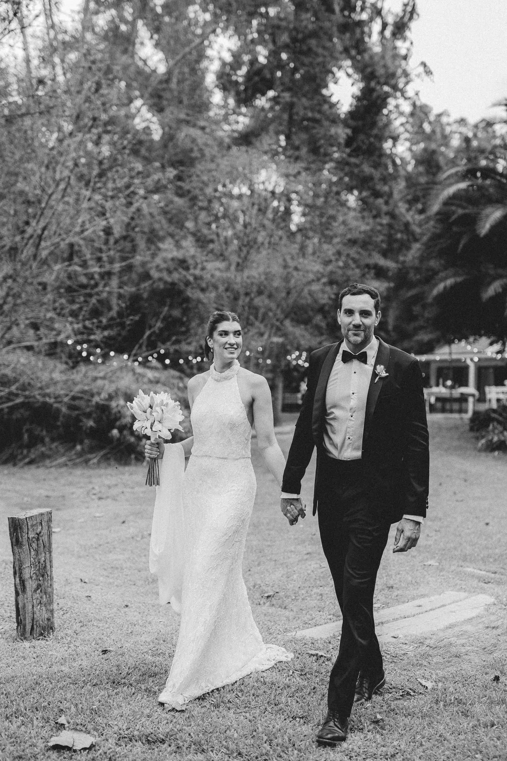 Los novios caminan de la mano por un parque arbolado, en blanco y negro.