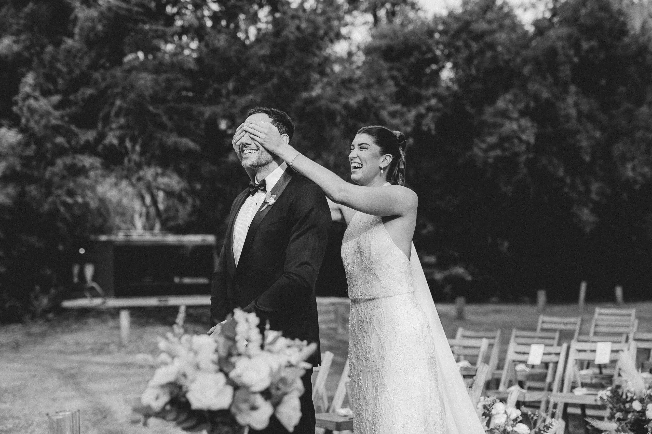 En blanco y negro, la novia le tapa los ojos al novio en el jardín, entre flores y las sillas de la ceremonia.